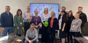 Image shows a group of training session attendees standing in front of a screen showing a snakes and ladders board. The training facilitator is holding an over-sized dice. 
