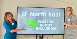 Image shows Lucy (a white woman wearing a blue dress) and Karen ( a white woman wearing glasses and a patterned dress) standing either side of a screen showing the Inclusion Forum North East logo. Both of them are smiling and excited.